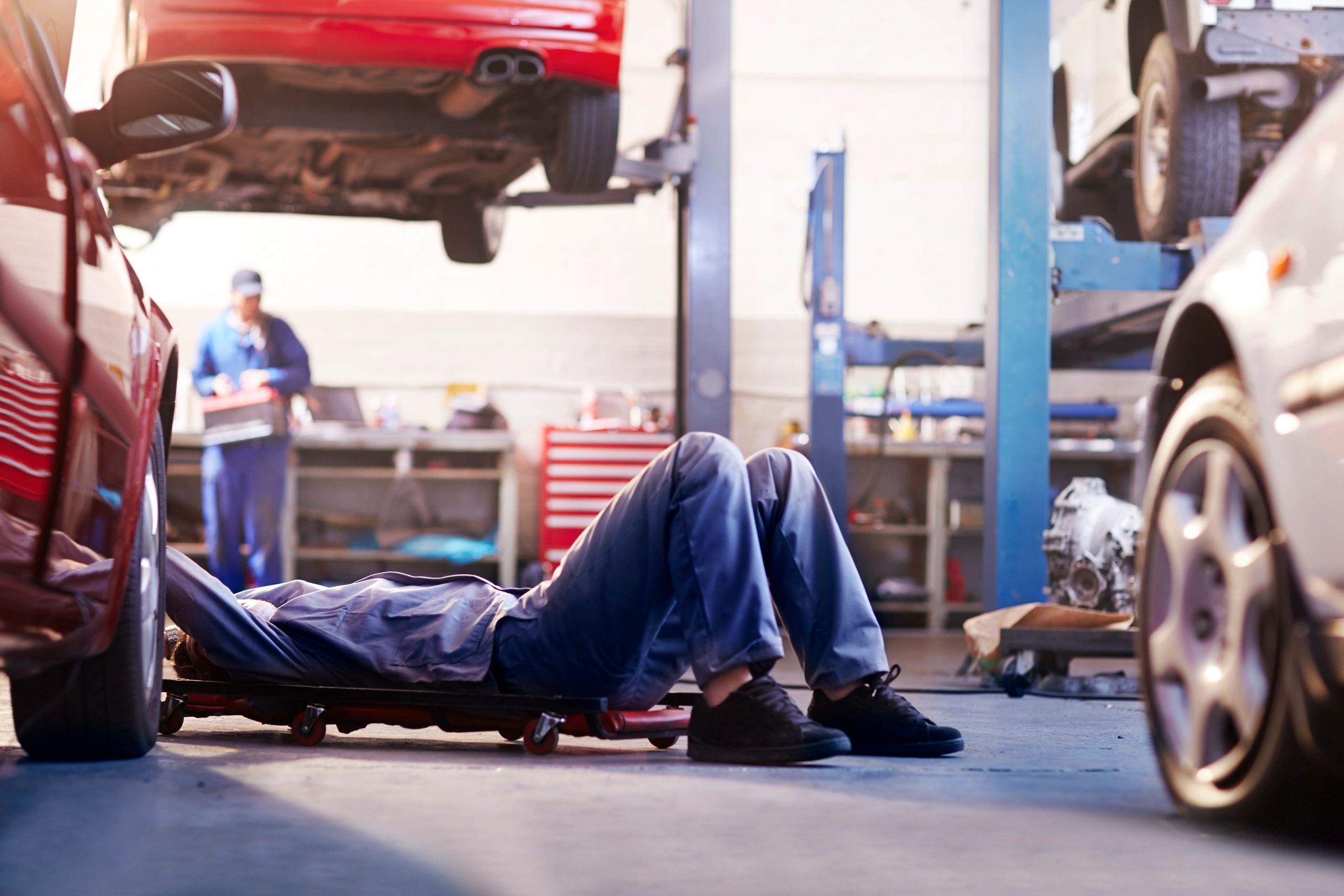 A mechanic working under a car in a garage, with other vehicles being serviced in the background.